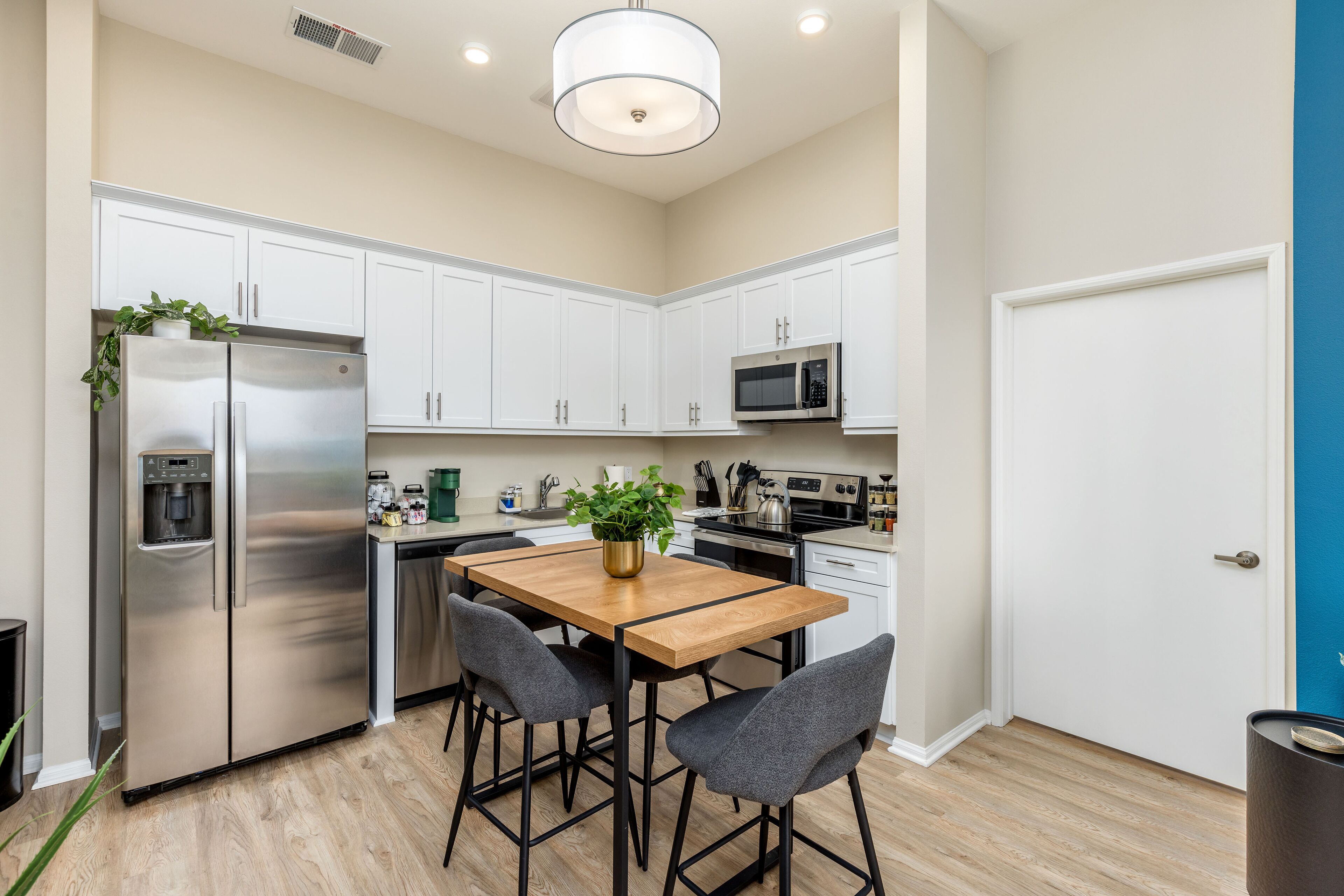 A compact kitchen with white cabinets, a stainless steel refrigerator, a microwave, and a wooden dining table with four chairs.