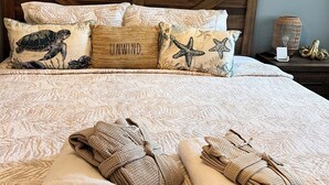 Ein ordentlich bezogenes Bett mit einem Strandkissen, einem Kopfteil und einem Gemälde mit Strandmotiv.