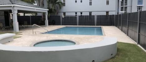 A swimming pool with a circular hot tub, surrounded by a fenced area with a covered seating area and palm trees.