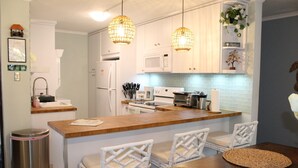 Una cocina moderna con armarios blancos, encimera de madera y dos sillas de mimbre. Hay lámparas colgantes de mimbre, un cubo de basura y un cartel que dice 'Coffee Time'.