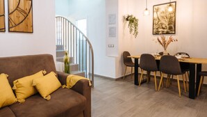 A modern living room with a brown sofa and yellow cushions, a dining area with a wooden table and chairs, and a staircase leading to an upper floor.