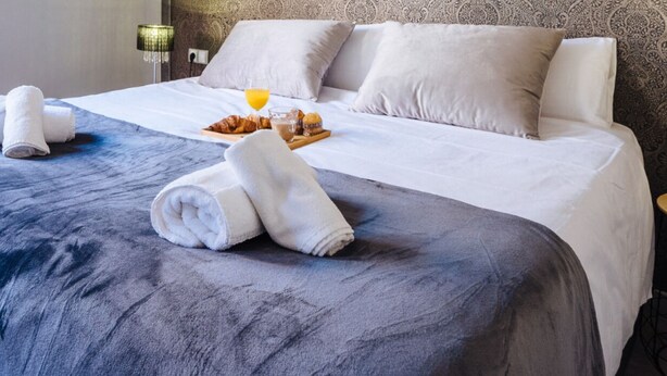 A neatly made bed with white linens, a tray of croissants and orange juice, and rolled towels on a blue-gray bedspread.