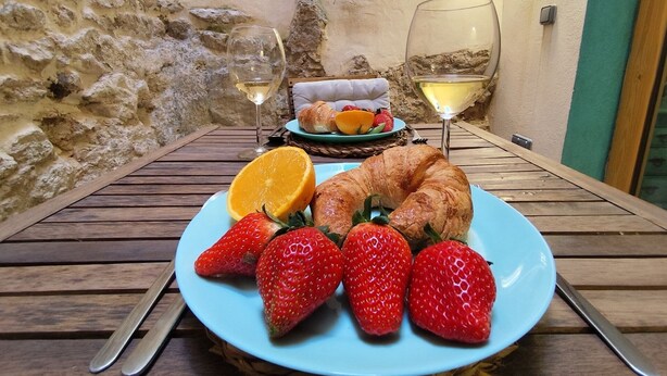 Une table dressée avec une assiette de fraises, une tranche d’orange et un croissant, accompagnée de deux verres de vin blanc.