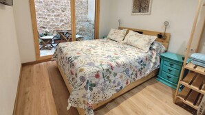 Une chambre avec un lit en bois, une couvre-lit à motifs floraux, une petite table avec des chaises, un mur en pierre et une vue par la fenêtre.