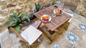 Une table en bois, dressée avec des assiettes remplies de nourriture et des boissons, une chaise, et des plantes en pot, le tout dans un espace extérieur dont les murs sont en pierre.