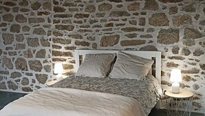 Ein Schlafzimmer mit Steinwand, einem Bett mit weißem Kopfteil, zwei Nachttischlampen und einem kleinen runden Tisch mit Lampe.