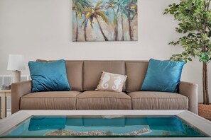 A living room with a beige sofa, two blue cushions, a white cushion with a seahorse design, a glass coffee table, a small indoor pool, a wall-mounted painting of palm trees, and a potted plant. - Cottage w/Heated Pool~10 Mins from Downtown &Beach (St. Augustine)
