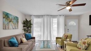 Ein Wohnzimmer mit braunem Sofa, einem grünen Sessel, einem Couchtisch mit Glasplatte und einem Deckenventilator.