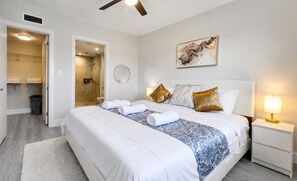 A hotel room with a bed, a bedside table, a nightstand, a mirror, a shower, and a bathroom.