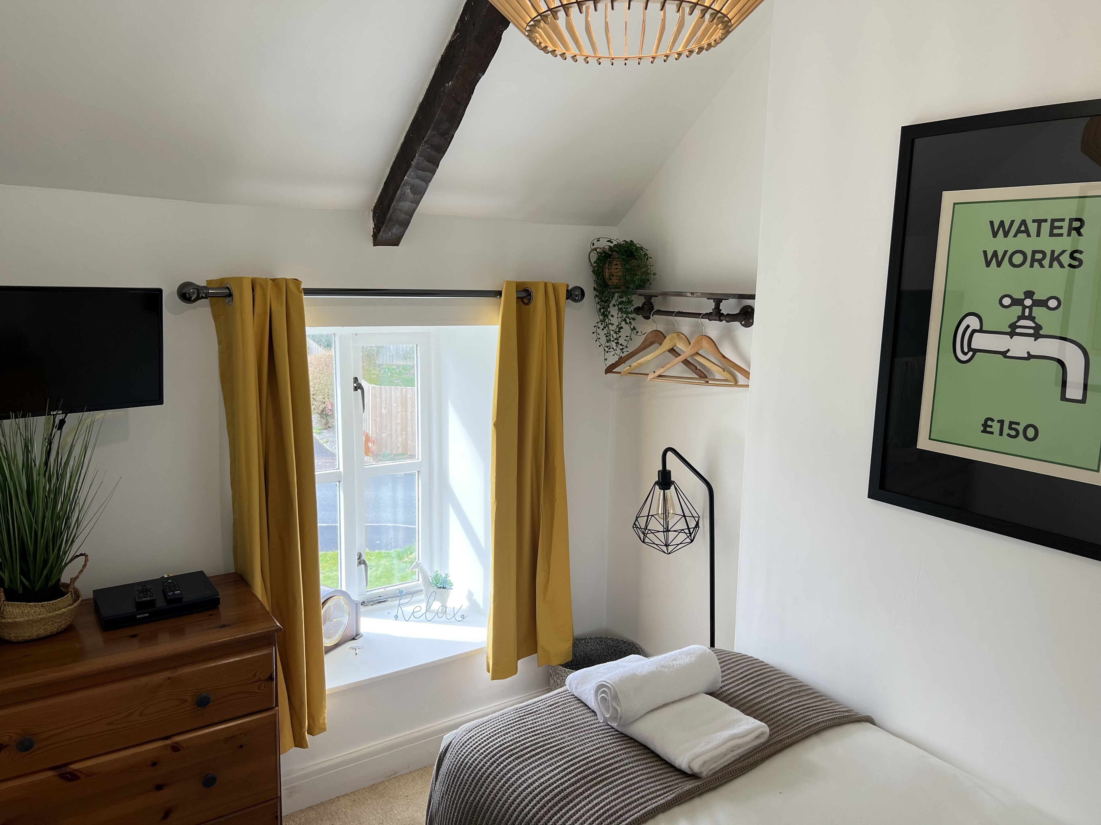 A bedroom with a bed, a bedside table with a TV, a window with curtains, a wall-mounted plant, a framed picture, and a towel on the bed.