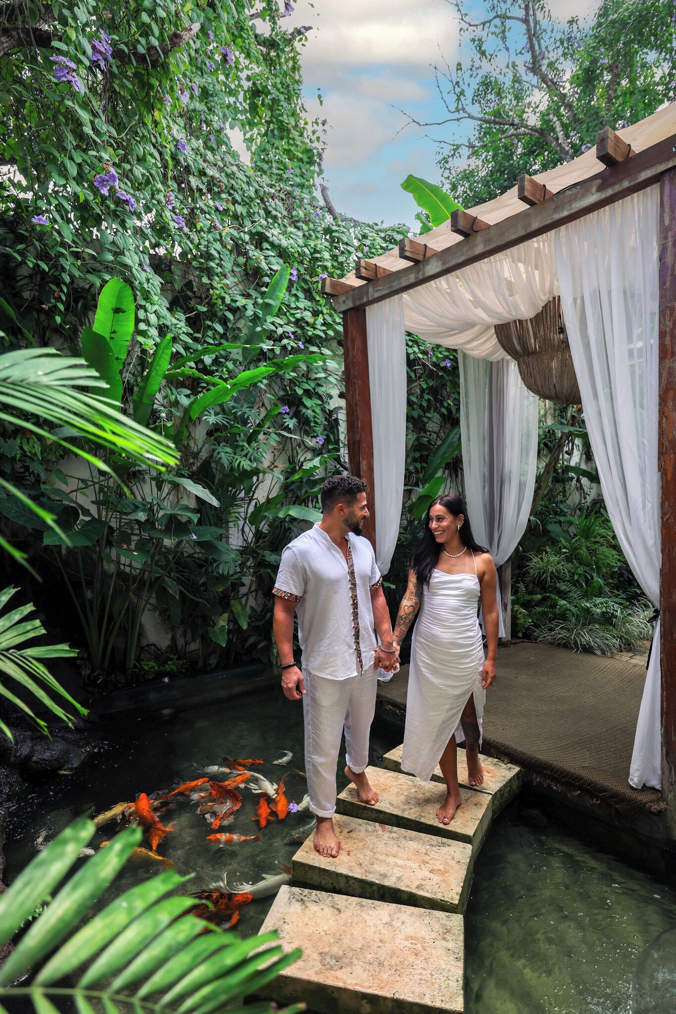 A couple standing on stone steps in a lush garden with a koi pond.