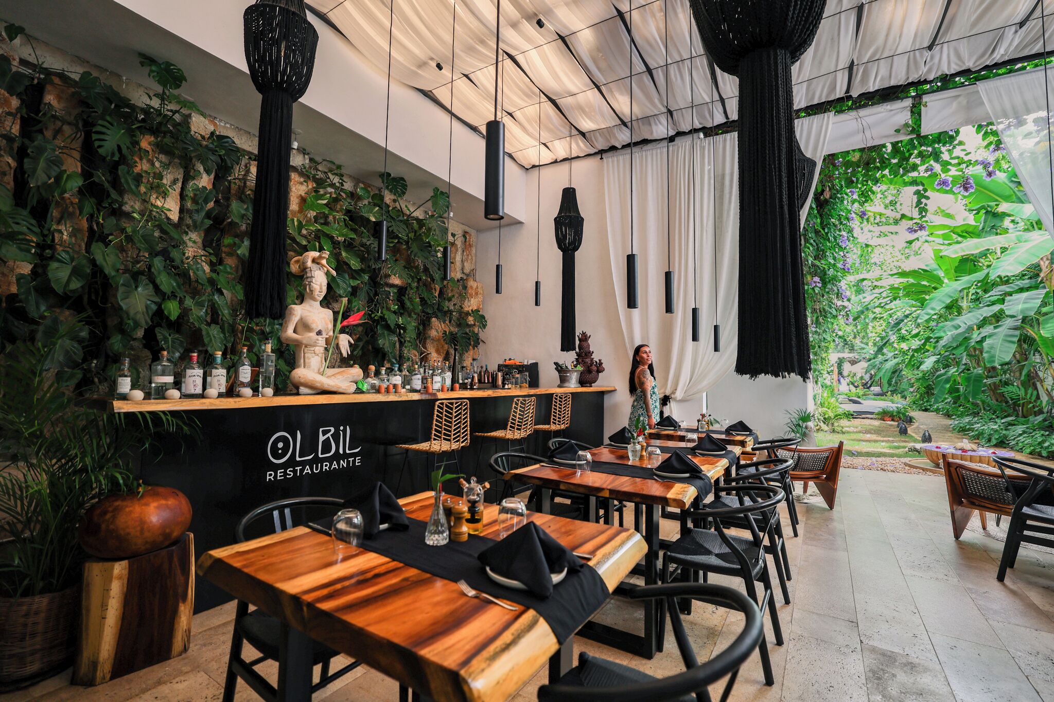 An indoor restaurant with wooden tables and chairs, a bar area with bottles, and a view of a lush garden through a large window.