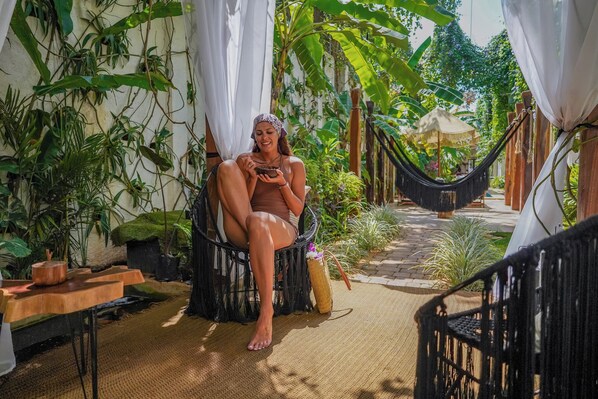 A person sitting in a hammock in a lush, green outdoor area with plants and a wooden table.