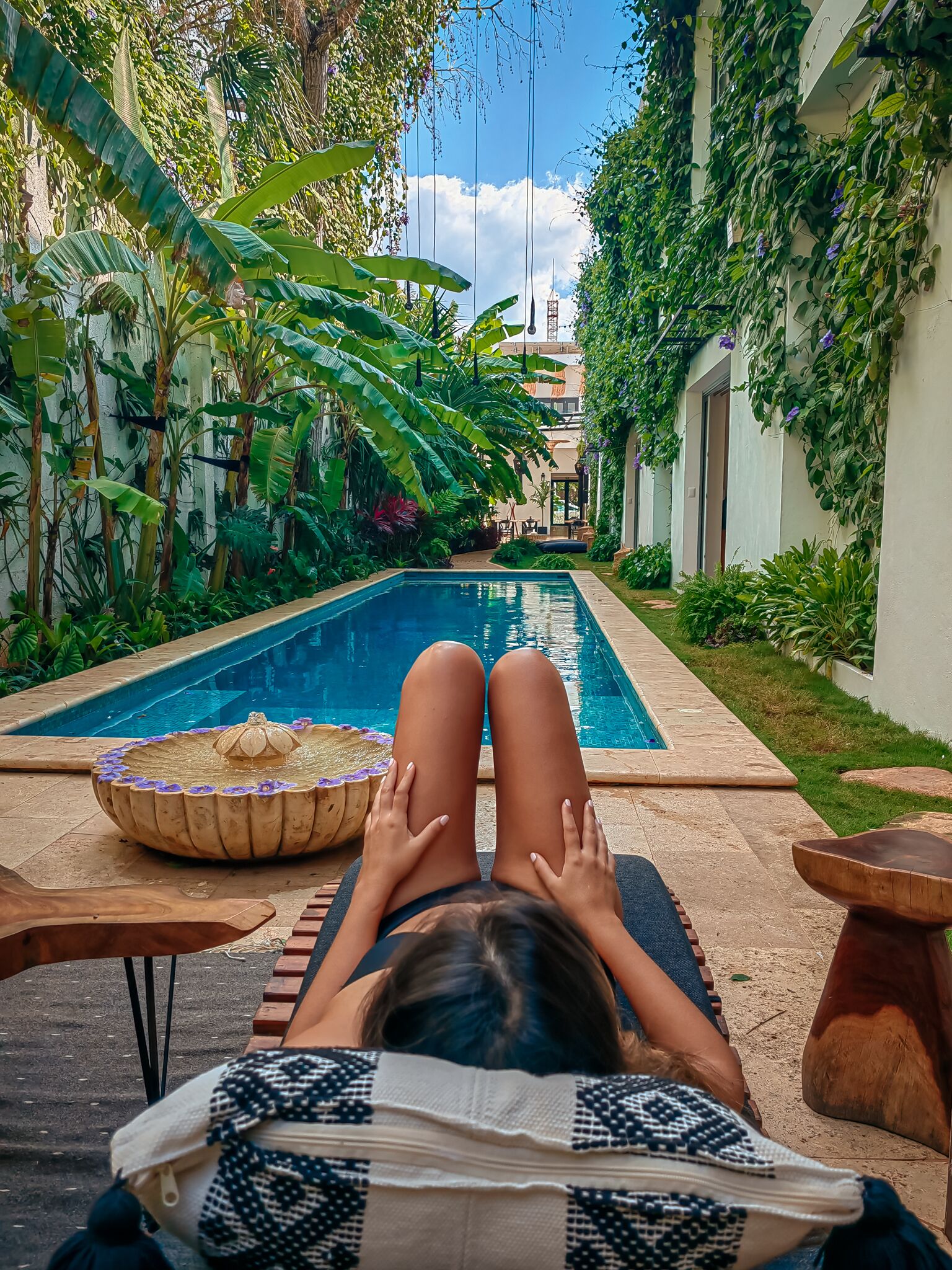 Zona junto a la piscina con una persona relajándose en una tumbona, rodeada de exuberante vegetación y una piscina de color azul cristalino.
