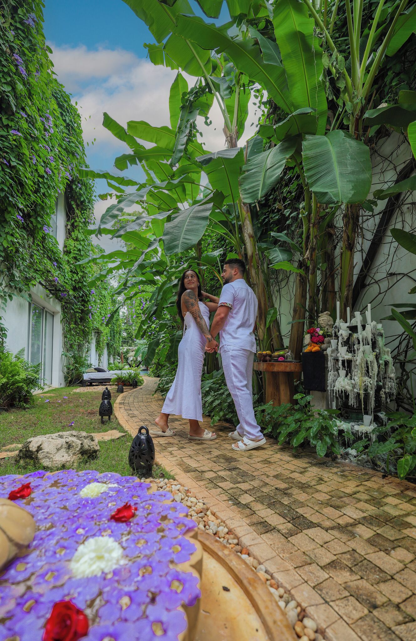 Una pareja tomándose de las manos en un exuberante jardín con palmeras de plátano y un sendero de piedra.