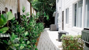 Un étroit sentier pavé de briques, bordé de plantes verdoyantes et d’un bâtiment blanc doté de grandes fenêtres.