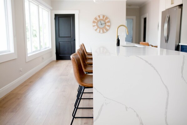 A modern kitchen with a marble countertop, wooden chairs, and a clock on the wall.
