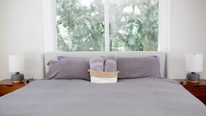 Un lit bien fait, avec une couverture grise, deux oreillers et un panier rempli de serviettes pliées.