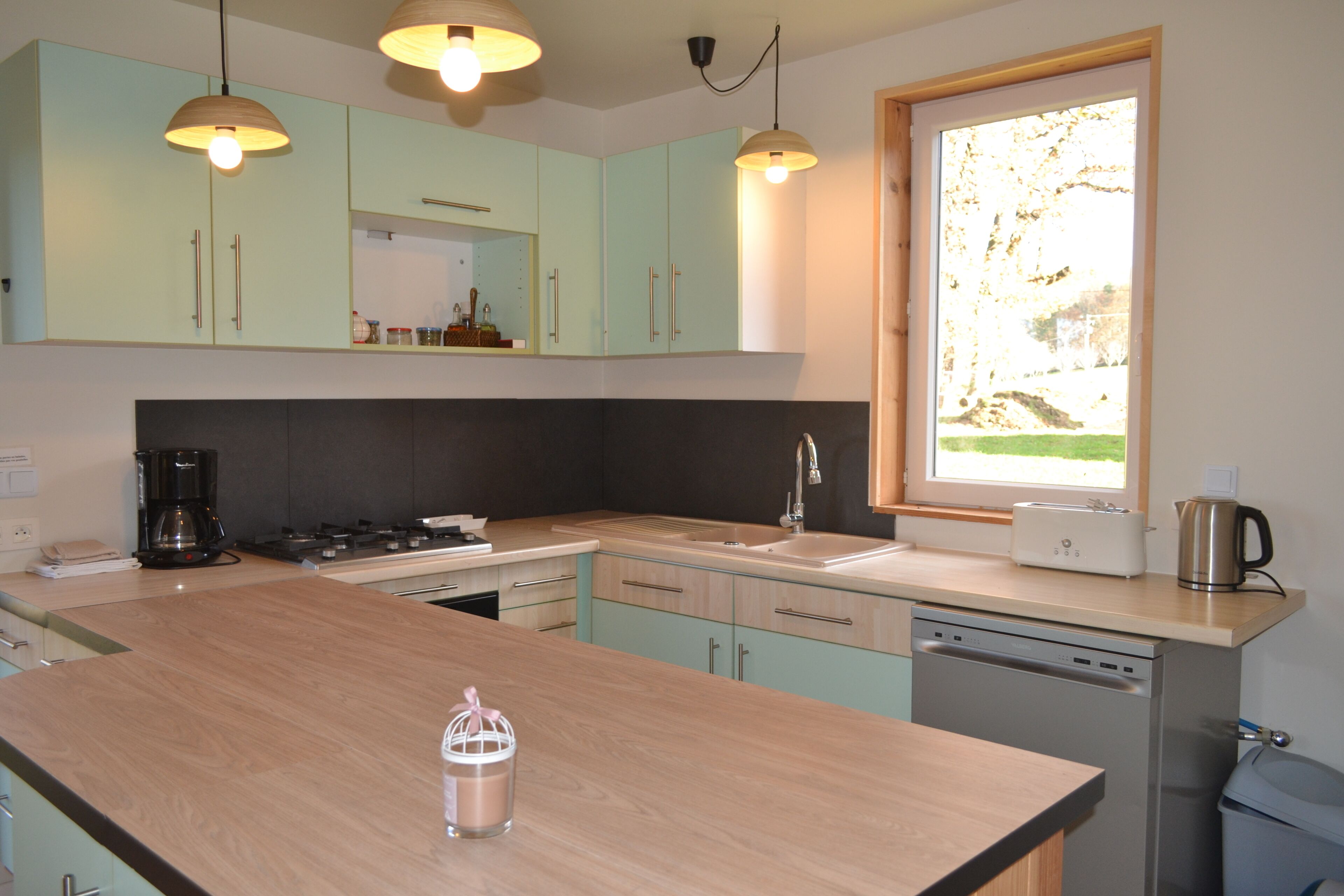 Een moderne keuken met lichtgroene kastjes, een houten aanrechtblad, een ingebouwde vaatwasser en hanglampen.