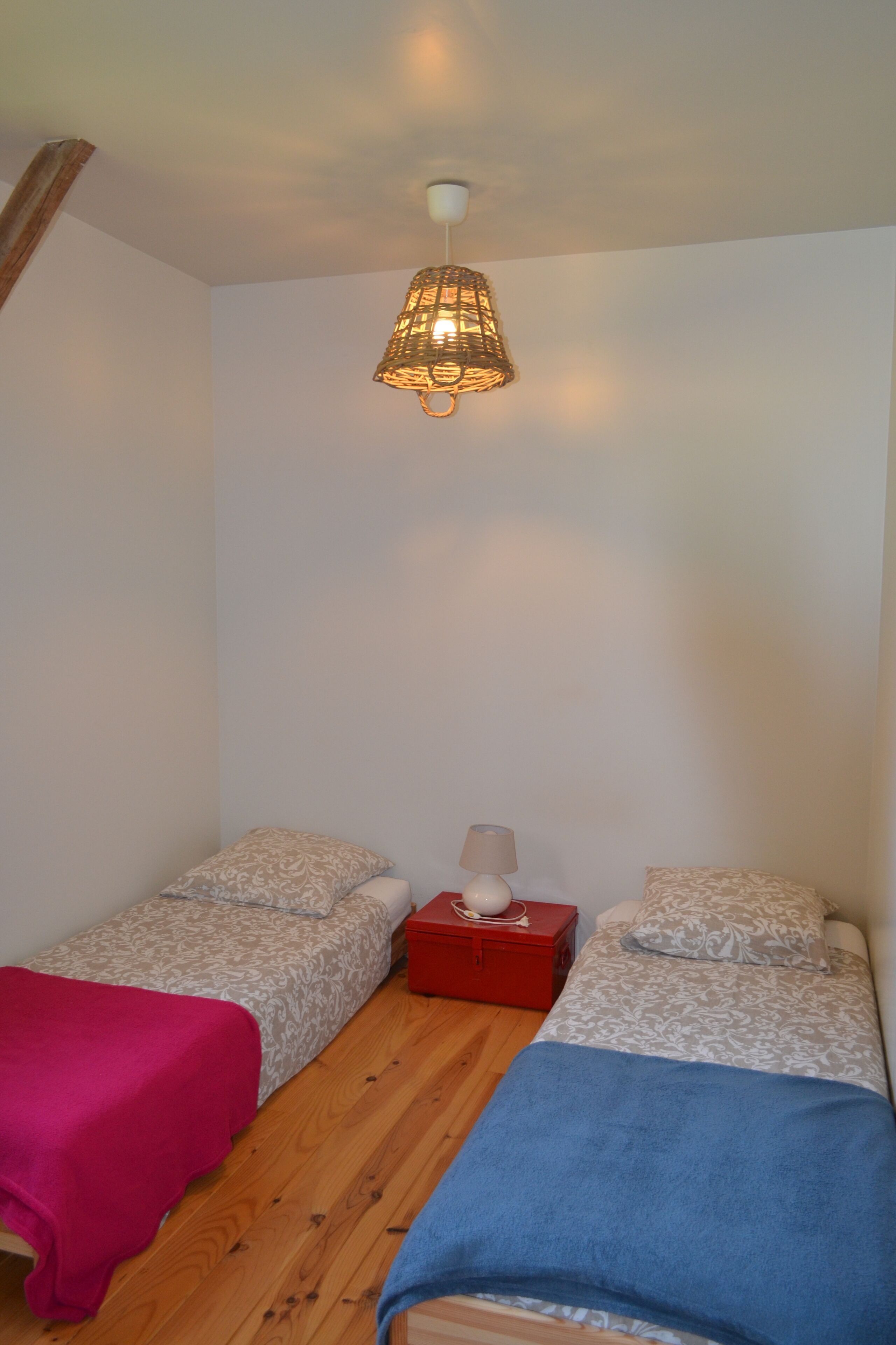 A small, single-room accommodation with two beds, a wooden floor, a red nightstand, and a hanging light fixture.