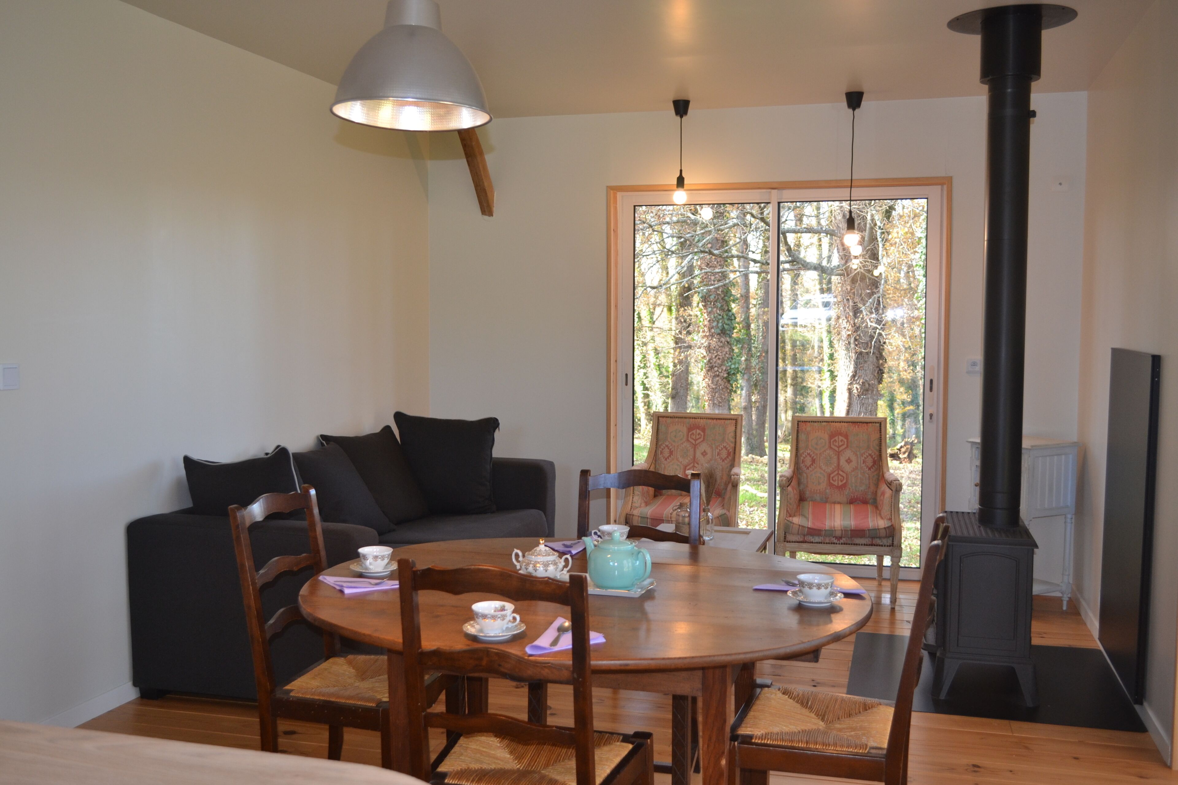 Een gezellige woonkamer met een ronde houten tafel, stoelen, een bank en een groot raam met uitzicht op bomen.