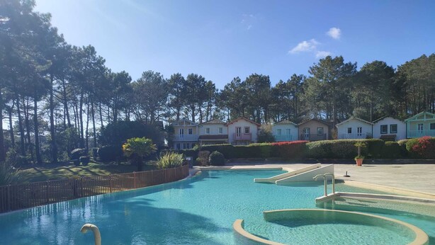 A swimming pool with a clear blue water feature, surrounded by a wooden fence and a row of colorful houses in the background. - La maisonnette du Golf (Lacanau)