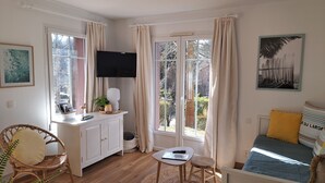 A cozy living room with a wooden floor, a white cabinet, a small round table, a wicker chair, a sofa with cushions, and a television on the wall. - La maisonnette du Golf (Lacanau)