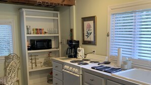 A kitchen with white cabinets, a stove, a microwave, and a coffee maker. There is a window with blinds and a chair with a cushion.