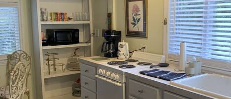 A kitchen with white cabinets, a stove, a microwave, and a coffee maker. There is a window with blinds and a chair with a cushion.