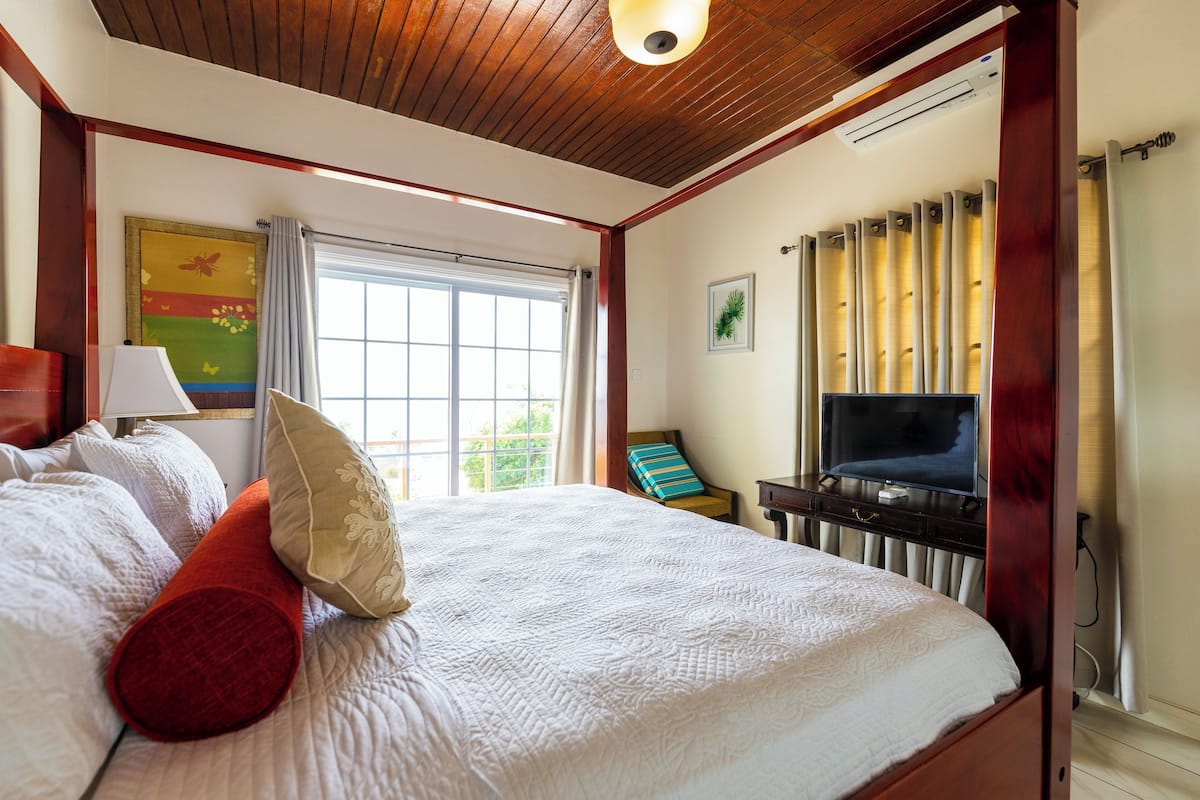 A bedroom with a large bed, a TV, a window with curtains, and a painting on the wall.