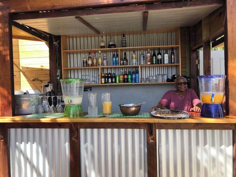 Een bar met een houten toonbank, planken met verschillende flessen, een blender en een persoon achter de toonbank.