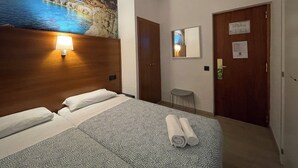 Habitación de hotel con cama, mesita de noche con lámpara, espejo en la pared y puerta con un cartel.