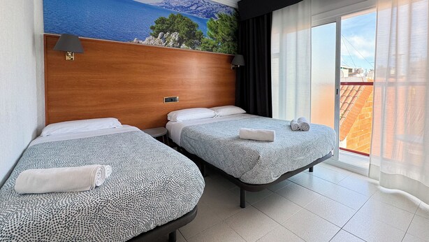 Habitación de hotel con dos camas, cabecera de madera, un mural con paisaje costero y vistas a edificios a través de cortinas translúcidas.