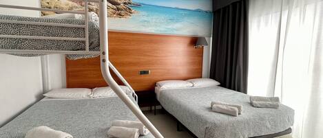 A hotel room with bunk beds, two single beds, a wooden headboard, a nightstand, a lamp, and a beach-themed wall mural.