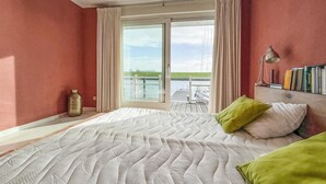 Een slaapkamer met een groot bed, uitzicht op een steiger en water, en een boekenkast met boeken.