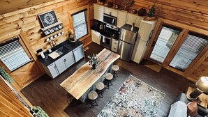 A cozy cabin-style kitchen with wooden walls, a dining area with stools, and a living room with a sofa and a coffee table.