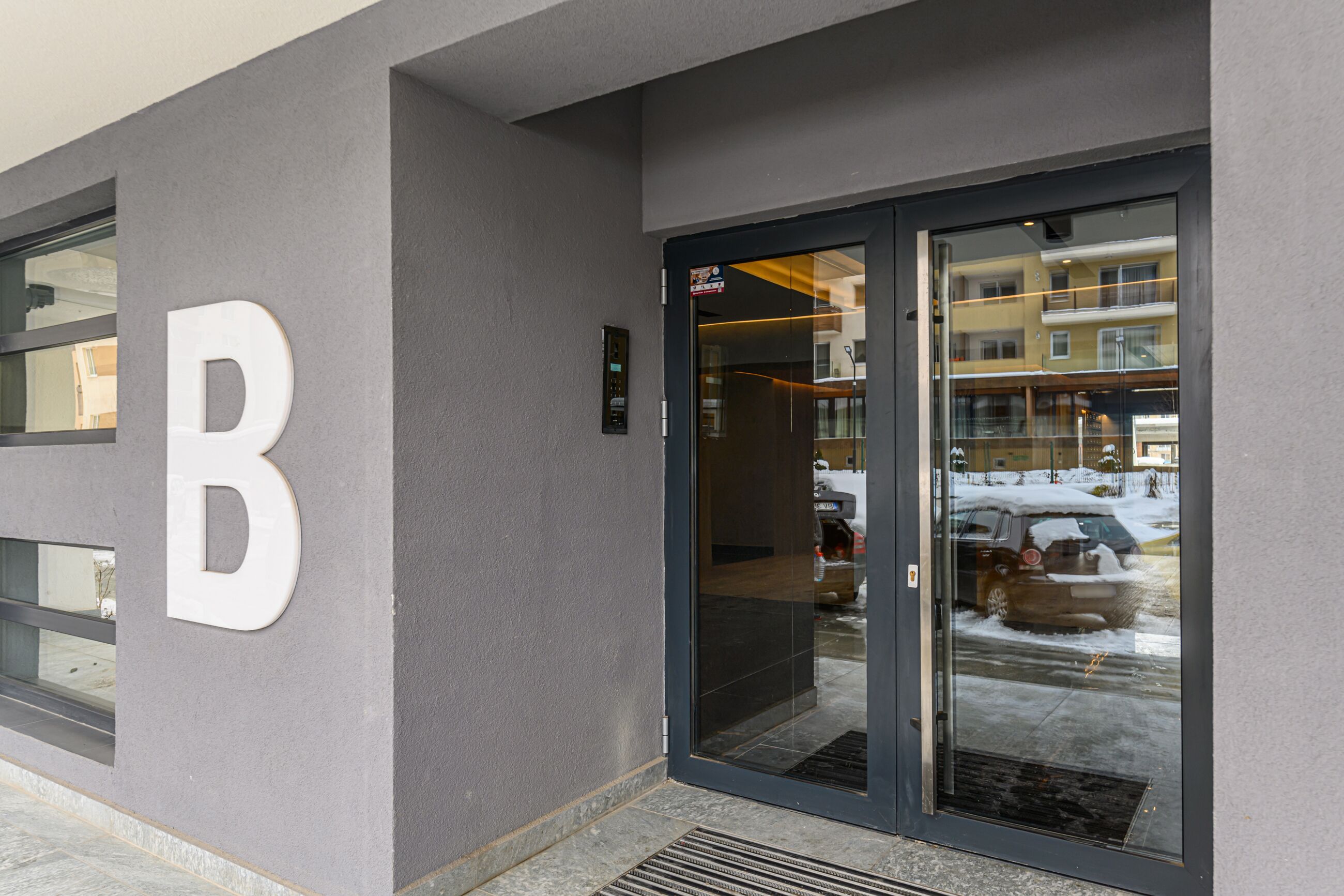 A building entrance with a large 'B' sign, a glass door, and a reflection of a snowy street with parked cars.