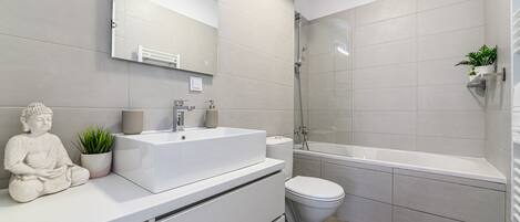 Un baño moderno con lavamanos, inodoro y bañera blancos. Hay un espejo grande sobre el lavamanos y una estatua de Buda en la encimera.