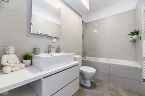 A modern bathroom with a white sink, toilet, and bathtub. There is a large mirror above the sink and a Buddha statue on the counter. - Nubia Apartment (Brasov)