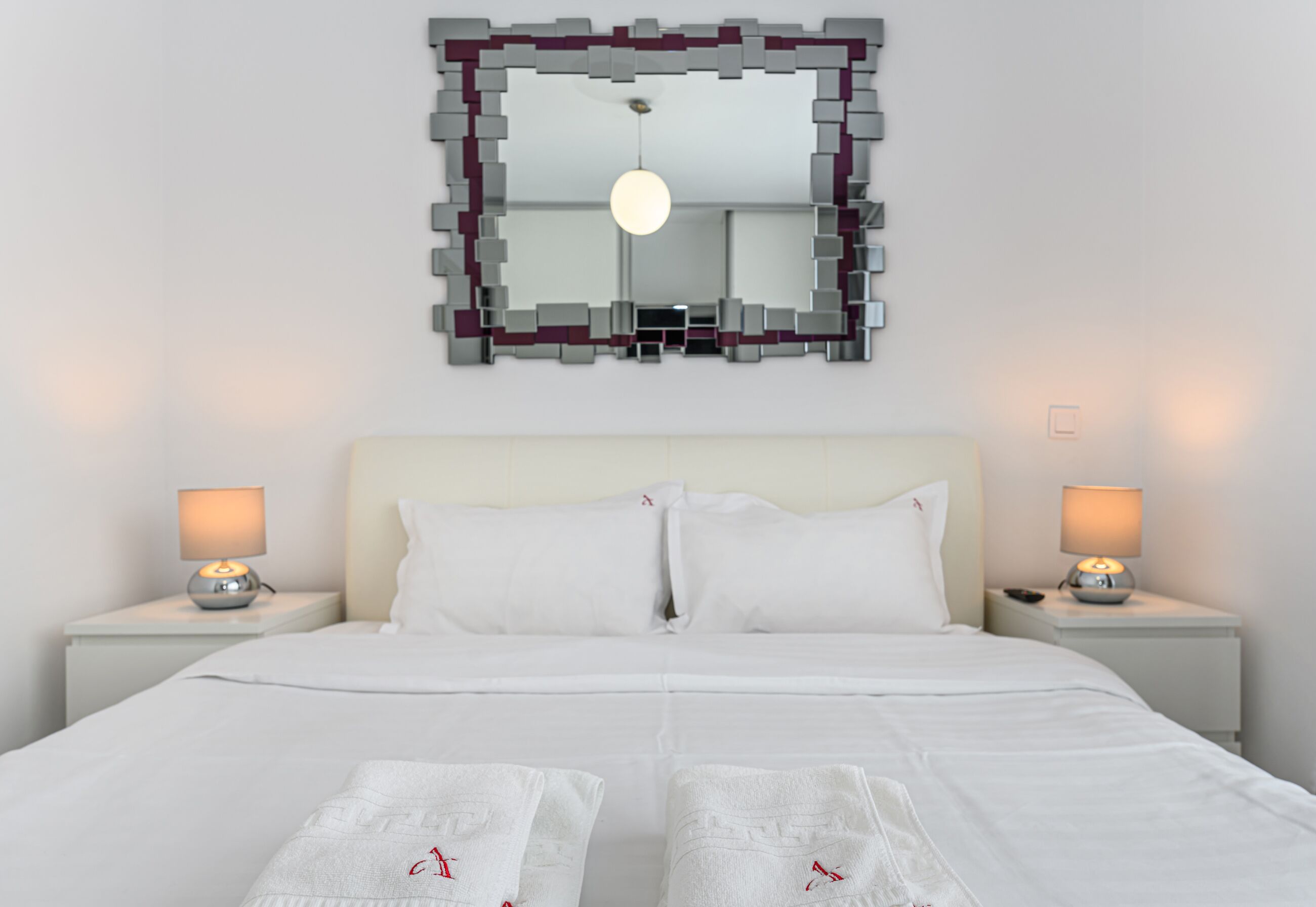 Un lit bien fait, avec du linge de lit blanc et des oreillers, deux lampes de chevet et un grand miroir doté d’un cadre géométrique.