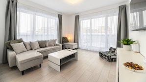 Ein modernes Wohnzimmer mit einer Couch, Hocker und Couchtisch, großen Fenstern mit durchscheinenden Vorhängen, einem kleinen Beistelltisch und einer Topfpflanze.