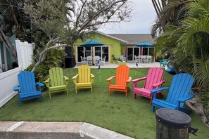 A backyard with a lawn, colorful Adirondack chairs, a barbecue grill, and a house with a patio. - The Flamingo Waterfront Bungalow (Madeira Beach)