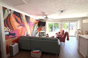 Una sala de estar con un mural colorido, un sofá gris, un ventilador de techo y vistas al exterior a través de un ventanal corredero.