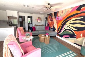 Ein modernes Wohnzimmer mit einem grauen Sofa, rosafarbenen Sesseln, einem Couchtisch und einem farbenfrohen abstrakten Wandbild.
