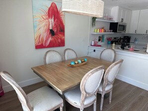 Un comedor con una mesa de madera y sillas blancas tapizadas, un cuadro de un flamenco en la pared, y una cocina al fondo.