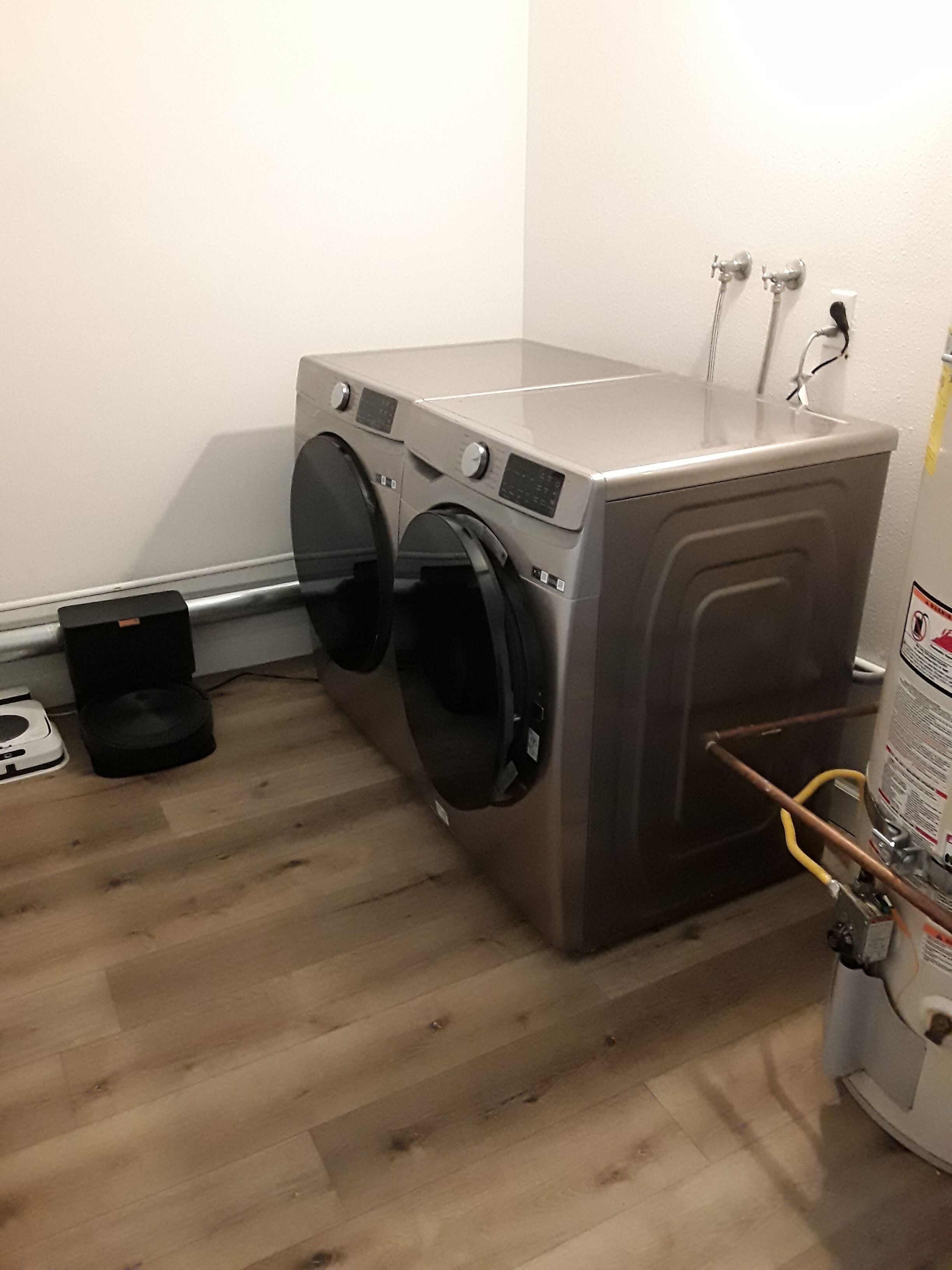 A compact laundry room with a front-loading washing machine and dryer, a small trash bin, and a water heater.
