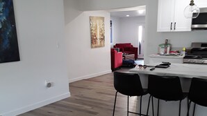 A modern living room with a wooden floor, a black sofa, and a red armchair. There is a kitchen area with a white countertop and bar stools. - Located in the heart of Palm Desert (Palm Desert)