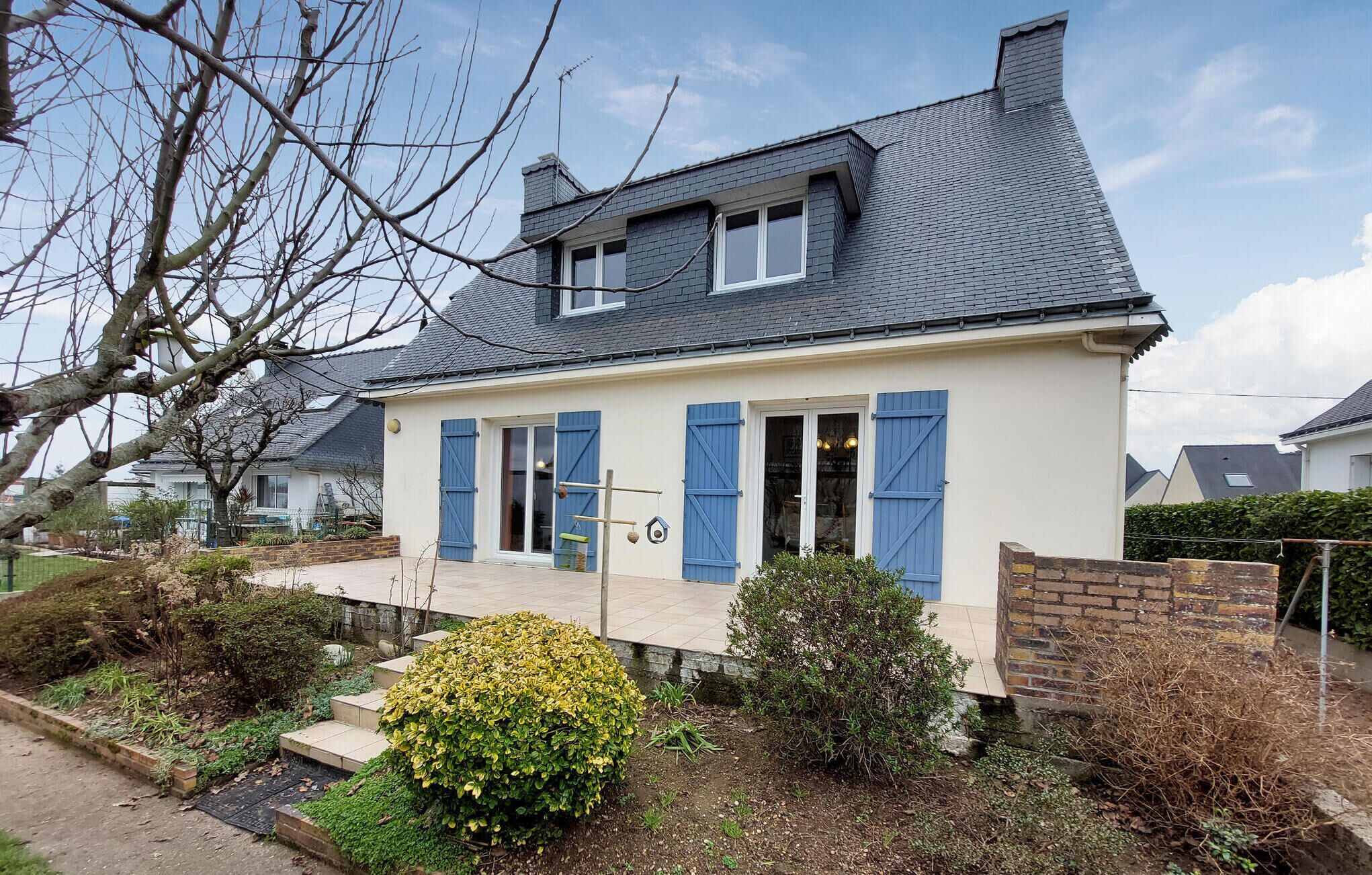 Een huis met twee verdiepingen, een donker dak, blauwe luiken en een kleine tuin met struiken en een gele bloem.