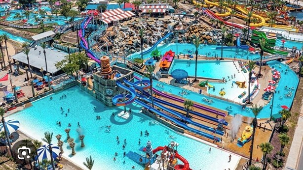Una vista aerea di un parco acquatico con diverse piscine, scivoli e un'ampia area piscina centrale.