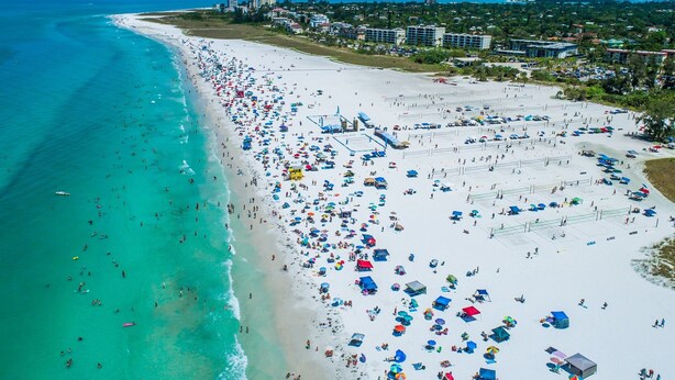 Eine Vogelperspektive zeigt einen Strand mit vielen Sonnenschirmen und Menschen, umgeben von Gebäuden und blauem Himmel.